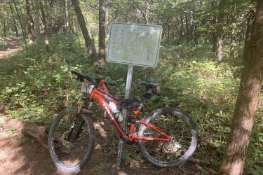 A mountain bike parked beside a trail sign in a wooded area, with lush green vegetation and trees in the background. The sign features a map of the trail and nearby routes. Cedar Niles Park mountain bike trail.