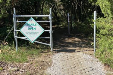 A metal gate with a "Trails Open" sign, indicating access to walking paths. The background features a dirt trail entering a wooded area, with greenery and trees on either side. There are additional signs visible along the trail. Cedar Niles Park mountain bike trail.
