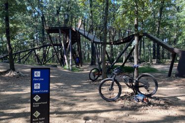 A forested area featuring a wooden bike trail structure in the background and two parked bicycles in the foreground. A directional sign points to various trail names, including "Oscar's Loop" and "Fire Line," with additional trail options listed below. The scene captures a sunny day with lush green trees surrounding the area. Coler Mountain Bike Preserve mountain bike trail.