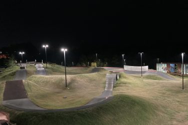 A pump track at night, illuminated by street lights. The track features smooth, winding paths and grassy mounds, with a wooden bridge and a barrier wall in the background. The surrounding area is dark, highlighting the track's lines and contours against the night sky. Railyard Bike Park mountain bike trail.