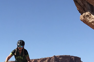A mountain biker navigating a rocky trail against a backdrop of red rock formations and a clear blue sky. The rider is wearing a black helmet and sunglasses, with yellow gloves and a black shirt featuring green graphics. North 40 mountain bike trail.