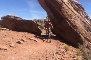 A mountain biker navigating a rugged dirt trail beneath a large, overhanging rock formation. The landscape features red earth and sparse vegetation under a clear blue sky. Lazy mountain bike trail.