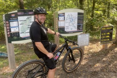 A smiling individual wearing a helmet is standing next to a mountain bike, leaning against it while pointing at a trail map. The background features dense trees and signage for mountain biking trails, including instructions and maps. The scene is set in a natural outdoor area, suggesting a hiking or biking destination. Little Sugar Creek mountain bike trail.