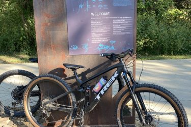 A black Trek mountain bike parked beside a large informational sign made of textured metal. The sign features illustrations and text detailing a design statement and welcoming visitors. Surrounding vegetation and a gravel path are visible in the background, indicating a natural setting suitable for biking. Coler Mountain Bike Preserve mountain bike trail.