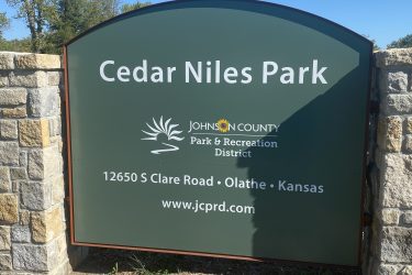 Sign for Cedar Niles Park, located at 12650 S Clare Road in Olathe, Kansas, under the jurisdiction of the Johnson County Park & Recreation District. The sign features a green background with white text and a stylized logo. Cedar Niles Park mountain bike trail.