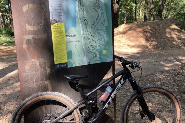 A mountain bike rests on a gravel path next to a trail map display in a wooded area. The map details various biking routes, including the North Gateway and Coler Camp, with color-coded trails and icons for different trail types. Sunlight filters through the trees, creating a vibrant outdoor setting. Coler Mountain Bike Preserve mountain bike trail.