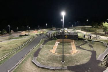 A nighttime view of a bike park featuring various dirt tracks and ramps, illuminated by overhead lights. The park has a winding paved path and wooden walkways, with several people engaging in recreational activities. The surrounding area includes grassy spaces and trees, contributing to a relaxed atmosphere. Railyard Bike Park mountain bike trail.