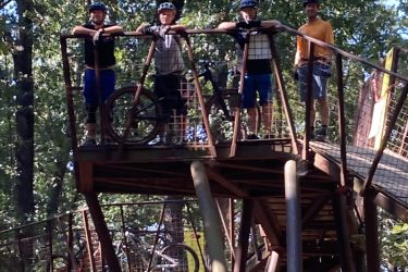 Four mountain bikers wearing helmets pose on a metal platform high above the ground, surrounded by tall trees in a wooded area. One biker holds a bike while the others lean on the railing, all smiling and enjoying the outdoor setting. The scene is bright and showcases the natural surroundings. Coler Mountain Bike Preserve mountain bike trail.