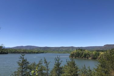 A serene landscape featuring a calm lake surrounded by lush green forests under a clear blue sky. In the distance, rolling hills and mountains create a picturesque backdrop, while a small boat can be seen gently gliding across the water. Lake James State Park mountain bike trail.