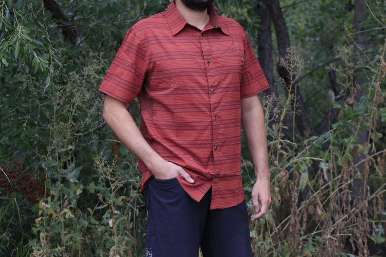A person standing outdoors in a natural setting, wearing a red and dark striped short-sleeve shirt and dark shorts. The background features greenery and tall grasses.