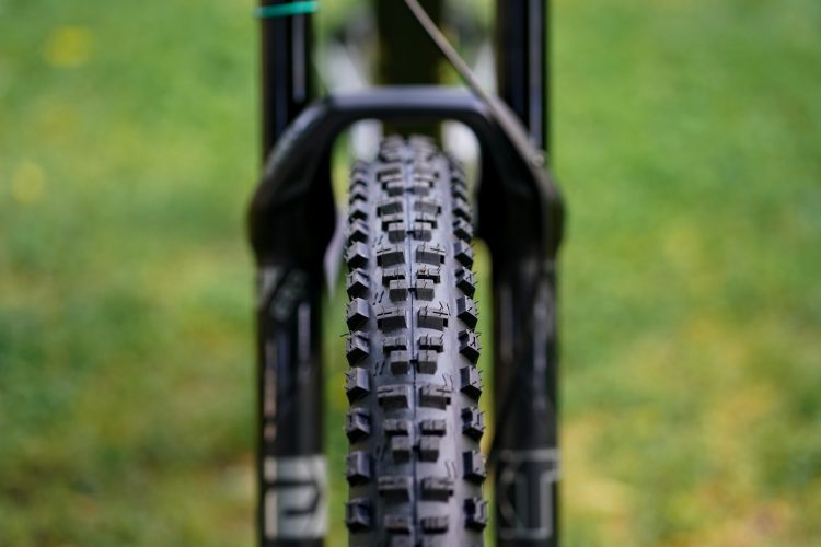 Close-up view of a bicycle tire with a rugged tread pattern, positioned between the forks of the bike. The background features a blurred green grass setting, creating a natural outdoor atmosphere.