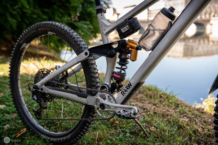 A close-up view of a mountain bike's rear suspension and drivetrain, showcasing the silver frame, black shock absorber, and detailed components. The bike is positioned on green grass near a body of water, with a water bottle attached to the frame. The background features blurred greenery and a bridge.