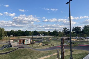 A panoramic view of a BMX and skate park featuring various ramps and hills, set against a backdrop of lush greenery and a blue sky with scattered clouds. A colorful shipping container serves as a unique structure in the park, while pathways wind through the grassy landscape. Several vehicles are parked nearby, indicating a community-friendly atmosphere. Railyard Bike Park mountain bike trail.