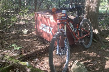 A mountain bike with an orange frame is parked beside a brown JOBBOX storage unit in a wooded area. The bike features a sign that reads "I'M RIDING TO FIGHT KIDS' CANCER" and is positioned on a dirt path surrounded by trees and underbrush. Sunlight filters through the foliage, creating a natural setting. Cedar Niles Park mountain bike trail.