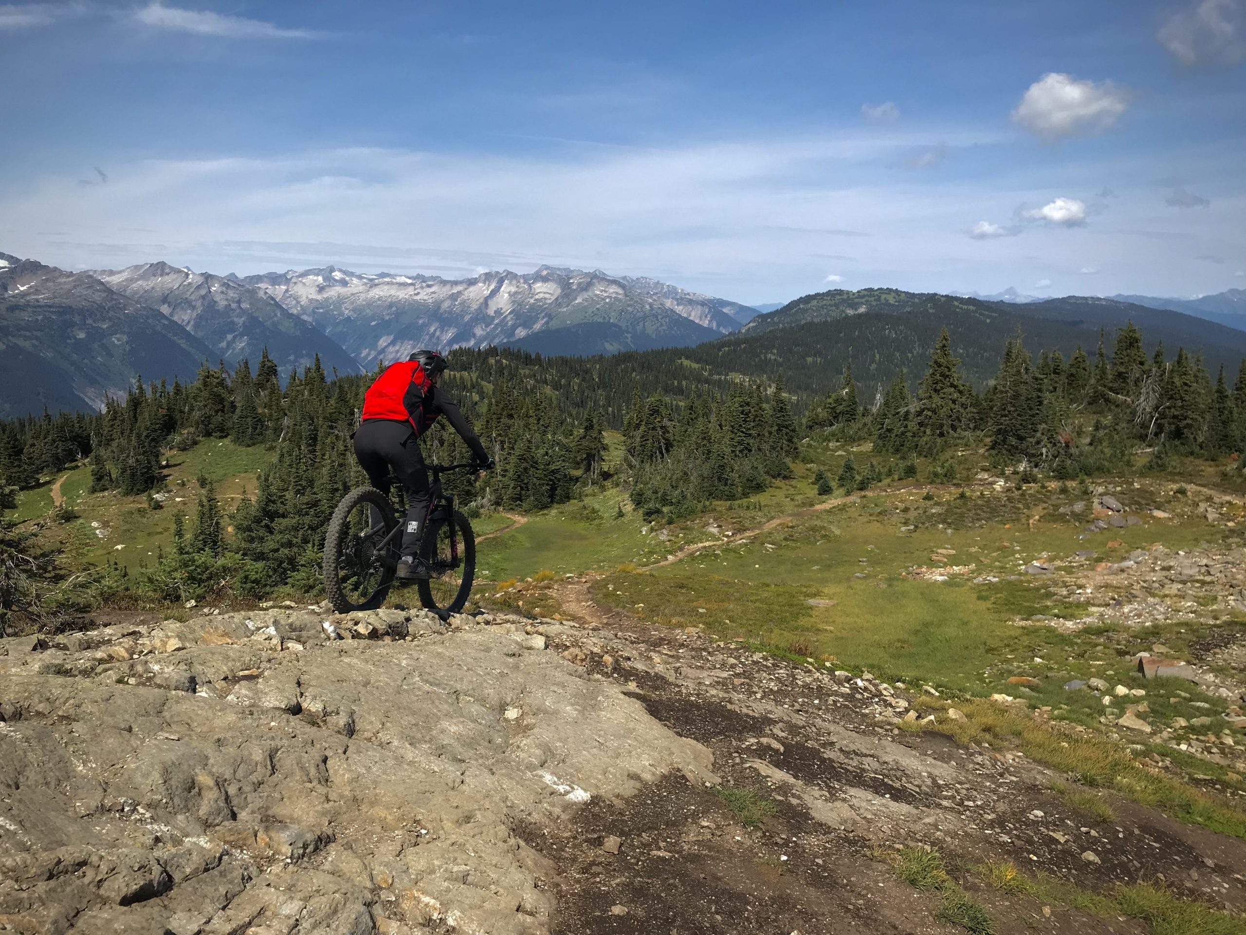 A mountain biker in a red jacket rides on rocky terrain overlooking a mountainous landscape. The scene features green pine trees and a winding trail, with snow-capped peaks visible in the background under a blue sky with scattered clouds. Frisby Ridge Trail mountain bike trail.