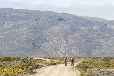 Three mountain bikers riding on a dirt trail through a rugged landscape with distant mountains and sparse vegetation under a partly cloudy sky. Anysberg Nature Reserve Trails mountain bike trail.