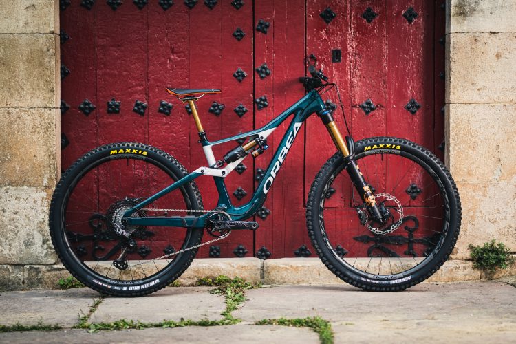 A mountain bike with a teal frame featuring white and orange accents is positioned against a rustic red wooden door adorned with black decorative elements. The bike has Maxxis tires and a high-performance suspension system, highlighting its rugged design suitable for off-road cycling. Green grass peeks through the stone pavement beneath the bike.