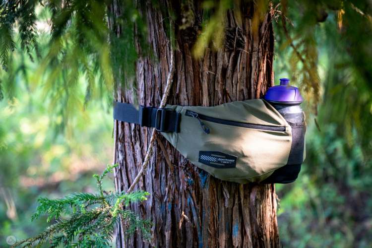 A sporty waist pack with a water bottle attached, hanging from a tree in a natural setting. The pack is olive green with a black zipper and logo, showcasing the bottle's purple lid. Surrounding foliage adds a vibrant green backdrop.