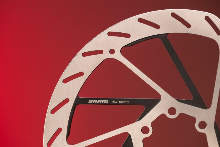 Close-up of a SRAM HS2 180mm bicycle brake disc, featuring a stainless steel surface with cutouts for weight reduction, set against a red gradient background.