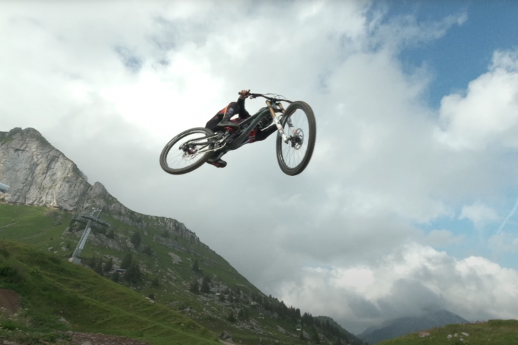 A mountain biker performing a mid-air jump over a grassy hillside with rocky cliffs and clouds in the background.
