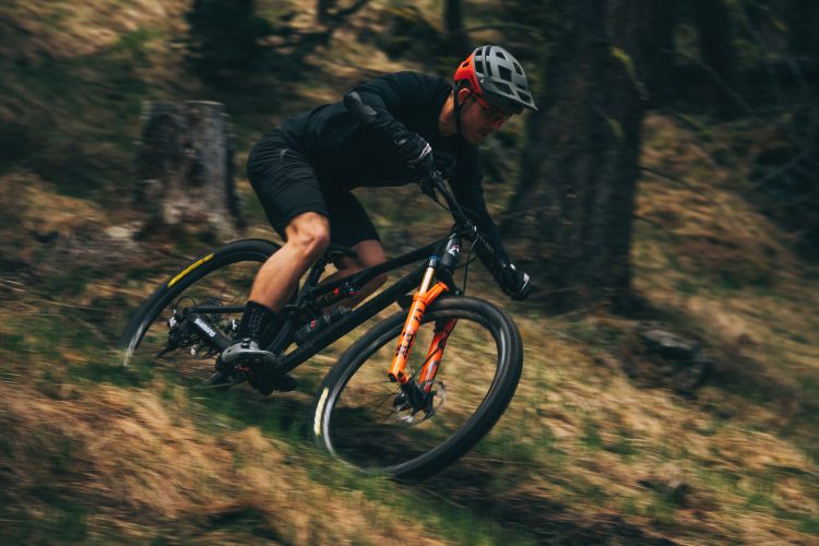 A cyclist in a black outfit and helmet rides a mountain bike down a grassy, forested trail, leaning into a turn. The scene captures the motion and excitement of mountain biking in a natural setting.