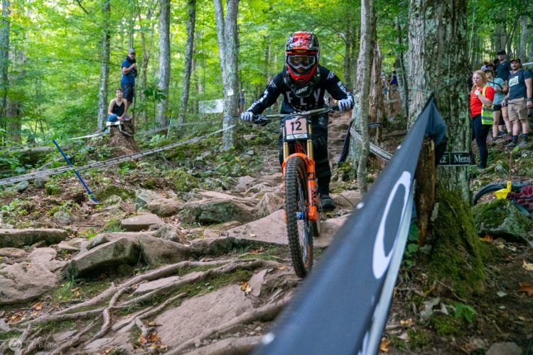 A mountain biker navigating a rocky trail in a forested area during a competition. The rider, wearing a helmet and protective gear, is focused on the challenging terrain, while spectators watch from the side. Trees and greenery surround the path, enhancing the outdoor racing atmosphere.