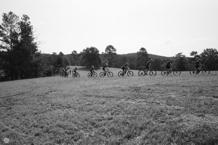 A group of mountain bikers riding in a line across a grassy field, set against a backdrop of trees and hills, captured in black and white.