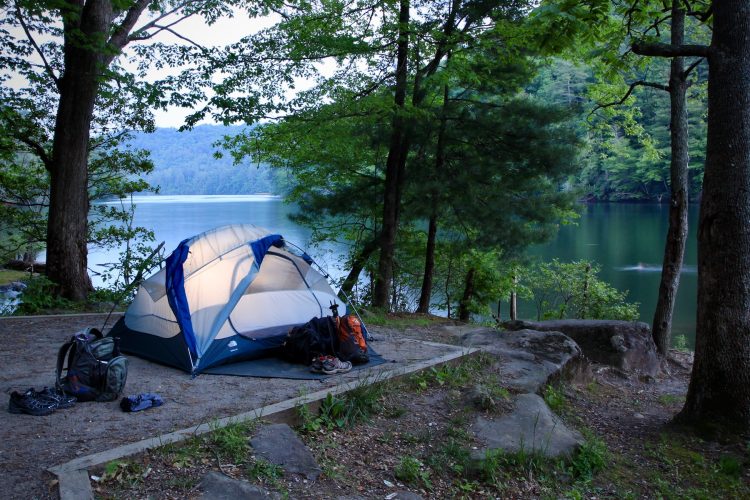 A tent set up by a calm lakeside, surrounded by lush greenery and trees. Backpacks and shoes are placed near the tent, with a serene body of water and distant mountains visible in the background under a twilight sky.