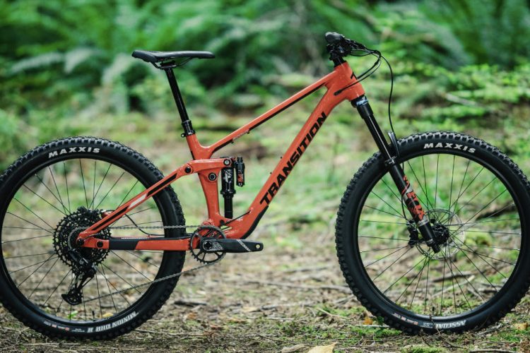 A bright orange mountain bike with a sleek design, featuring thick tires and a sturdy frame, positioned on a natural forest floor surrounded by greenery.