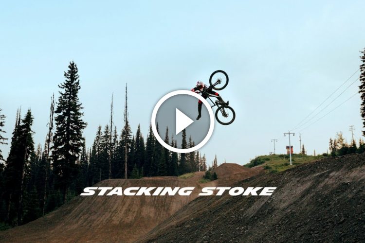 A mountain biker performs an impressive trick mid-air over a dirt jump, surrounded by tall evergreen trees under a clear sky. The text "STACKING STOKE" is prominently displayed at the bottom of the image.