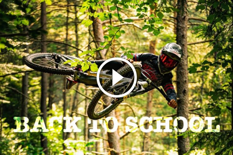A mountain biker performing a jump in a lush forest, surrounded by trees. The rider is wearing a helmet and protective gear, soaring through the air with their bike. The image features the text "BACK TO SCHOOL" prominently displayed at the bottom, alongside a play icon in the center.