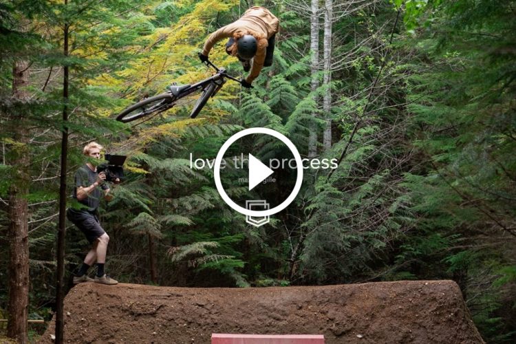 A mountain biker performs an airborne trick over a dirt ramp while a videographer captures the action in a forest setting, surrounded by lush greenery. The scene conveys excitement and adventure, emphasizing the connection between the rider and the outdoors.