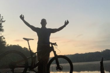 A silhouetted person stands with arms raised beside a bike at the edge of a serene lake during sunrise, surrounded by trees and reflecting water. Blankets Creek mountain bike trail.