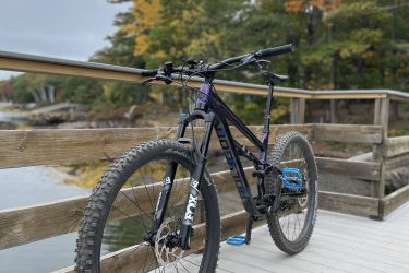 A mountain bike with blue pedal and bold, dark frame parked on a wooden bridge surrounded by autumn foliage and a calm waterway. The scene conveys a serene outdoor environment perfect for cycling. West Side Trail mountain bike trail.