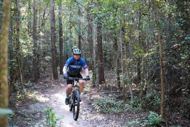 A person riding a mountain bike on a dirt trail surrounded by dense trees and greenery. The cyclist is wearing a helmet and a blue t-shirt with "Down East" written on it, navigating through a forested area. Big Branch Bike Park mountain bike trail.