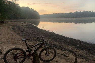 A mountain bike rests on the sandy shore of a tranquil lake at sunrise, surrounded by trees. The calm water reflects the soft hues of the early morning sky, creating a serene and peaceful atmosphere. Blankets Creek mountain bike trail.
