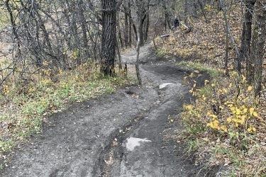 A winding dirt path leads through a wooded area with bare trees and patches of yellow foliage. The trail is slightly muddy and shows signs of use, while the surrounding landscape features rolling hills in the background. Lake Minnewasta Recreation Trail mountain bike trail.