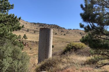 A solitary, cylindrical silo stands amid a grassy landscape, framed by pine trees. In the background, rolling hills are dotted with patches of evergreen trees under a clear blue sky. Picture Rock Trail mountain bike trail.