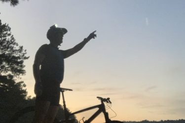 A silhouette of a person wearing a helmet, standing beside a mountain bike at dusk, pointing towards the sky. The scene includes a calm lake in the background and trees framing the edges, creating a tranquil outdoor atmosphere. The sky is transitioning from light to dark, with hints of color as the sun sets. Blankets Creek mountain bike trail.