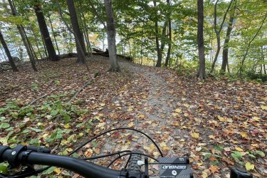 A view from the handlebars of a mountain bike, looking down a winding dirt path covered with colorful autumn leaves, surrounded by tall trees with green and yellow foliage. The scene suggests a peaceful ride through a forested area. West Side Trail mountain bike trail.
