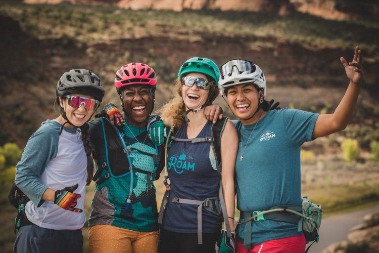 Four female mountain bikers, smiling and laughing, pose together in a scenic outdoor setting. They are wearing helmets and colorful biking gear, with a backdrop of rocky terrain and greenery. The atmosphere is joyful and friendly, showcasing camaraderie among the cyclists.