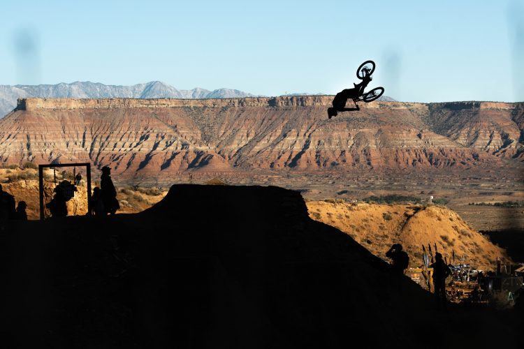 A silhouette of a cyclist performing a stunt in mid-air, against a backdrop of rugged mountains and clear blue sky. Spectators are visible in the foreground, observing the action in a desert landscape. The scene captures the thrill of extreme sports in a natural setting.