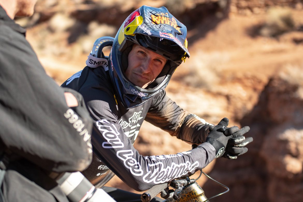 A mountain biker in a helmet and long-sleeve jersey sits on his bike, looking thoughtfully at a person off-camera. The background features rocky terrain and warm tones, suggesting an outdoor setting. The biker's gloves are clenched together, conveying a sense of focus or contemplation.