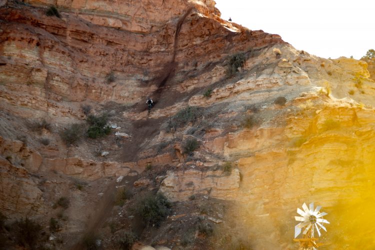A mountain biker descending a steep, rocky slope with layered red and orange stone cliffs in the background. A windmill is visible at the bottom of the slope, surrounded by sparse vegetation.