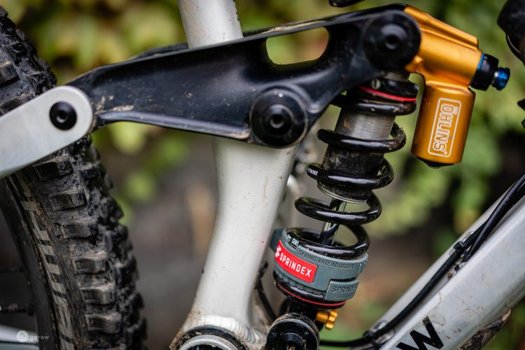 Close-up of a mountain bike's rear suspension system, featuring a black coil shock absorber with a red and gray spring rate adjuster labeled "SPRINDEX." The aluminum frame and components, including a black suspension linkage and yellow shock body, are visible alongside a muddy tire, hinting at off-road use. Green foliage in the background provides a natural setting.