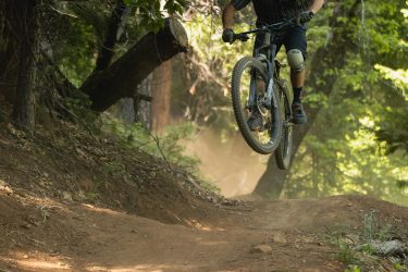 A mountain biker performing a jump on a dirt trail surrounded by trees, with dust flying up from the ground. The biker is wearing a helmet and protective gear, showing a dynamic action pose mid-air against a backdrop of lush green foliage. Hoot Trail mountain bike trail.