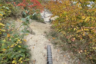 A mountain bike view from a narrow dirt path leading towards a serene lake surrounded by colorful autumn foliage, including vibrant red and yellow leaves. The cloudy sky adds a moody atmosphere to the picturesque landscape. West Side Trail mountain bike trail.