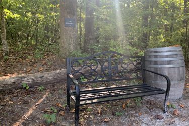A black metal bench beside a wooden barrel in a forested area, with a sign on a tree reading "THE HIDEAWAY." Sunlight filters through the trees, creating a serene and inviting atmosphere. Big Branch Bike Park mountain bike trail.