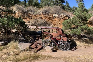 An abandoned, rusty vintage car sits against a rocky backdrop, surrounded by sparse vegetation and trees. In front of the car, two mountain bikes are parked on a rocky pathway, under clear blue skies. Picture Rock Trail mountain bike trail.