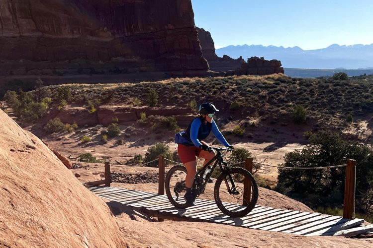 A mountain biker navigating a wooden path on rocky terrain, surrounded by red rock formations and vibrant desert vegetation. The background features distant mountains under a clear blue sky.